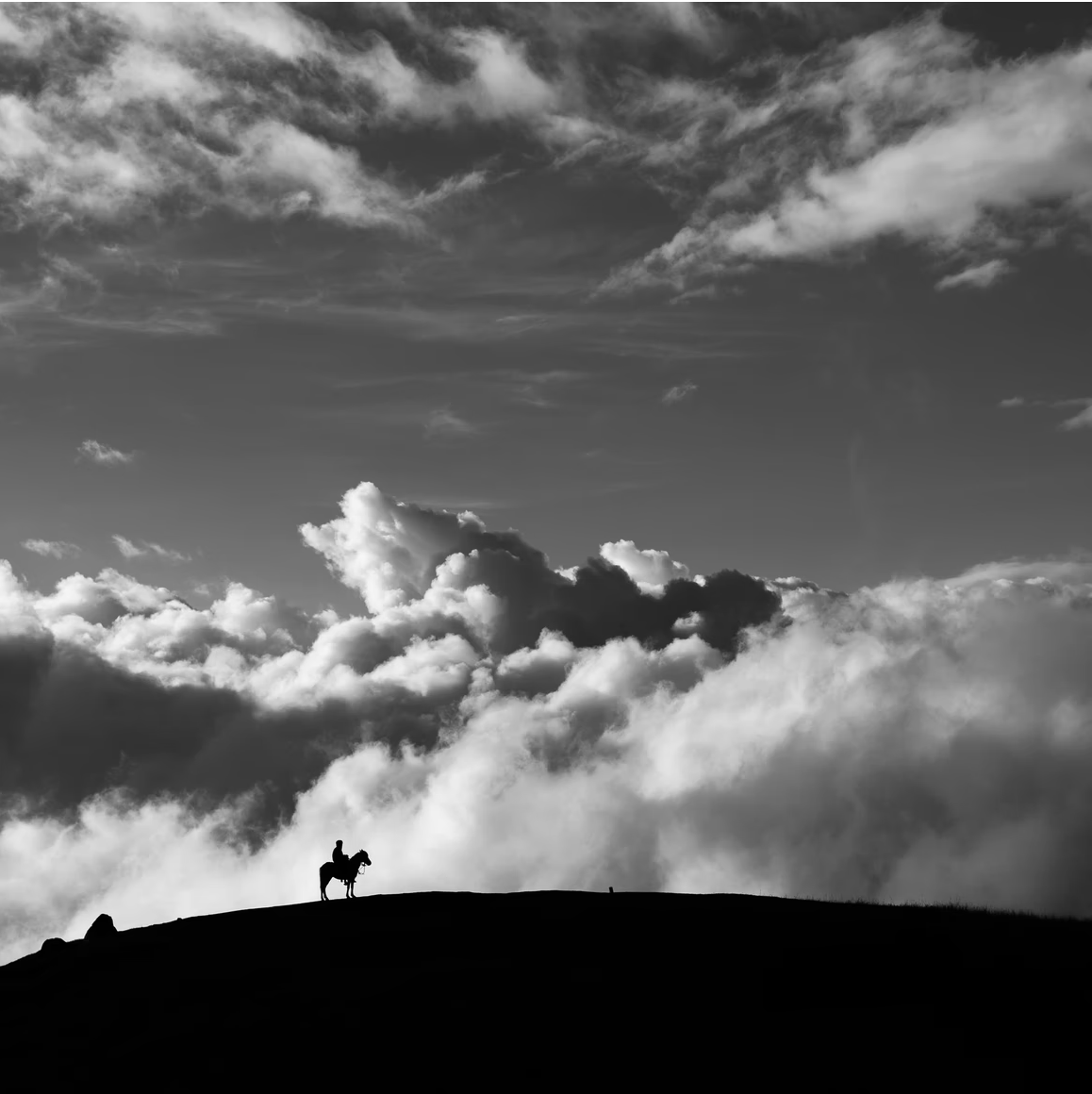 Rider on a high hill with clouds all around as he sits on his horse.