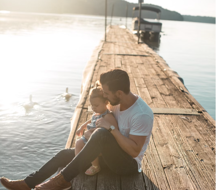 Young man with daughter on his lap on a pier jutting into a lake.