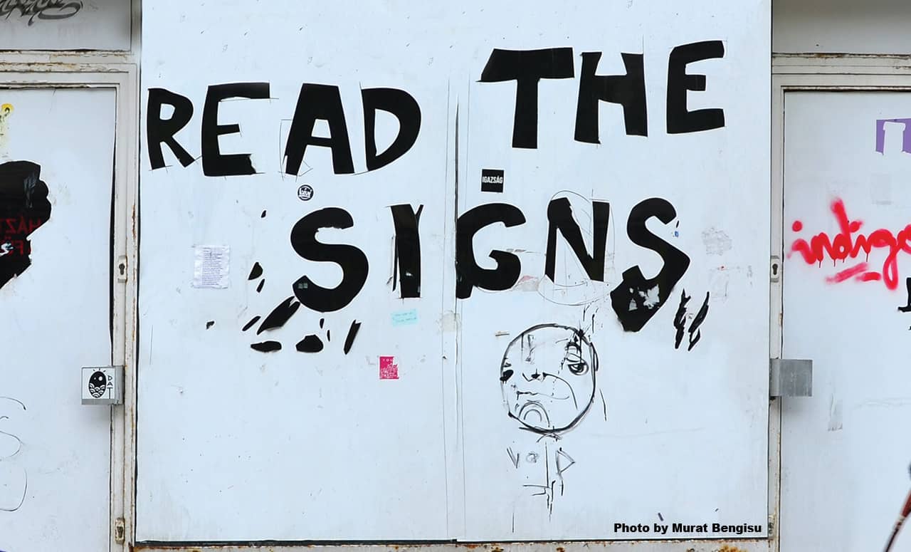 A photo of graffiti on the side of a building which reads, "Read the signs"
