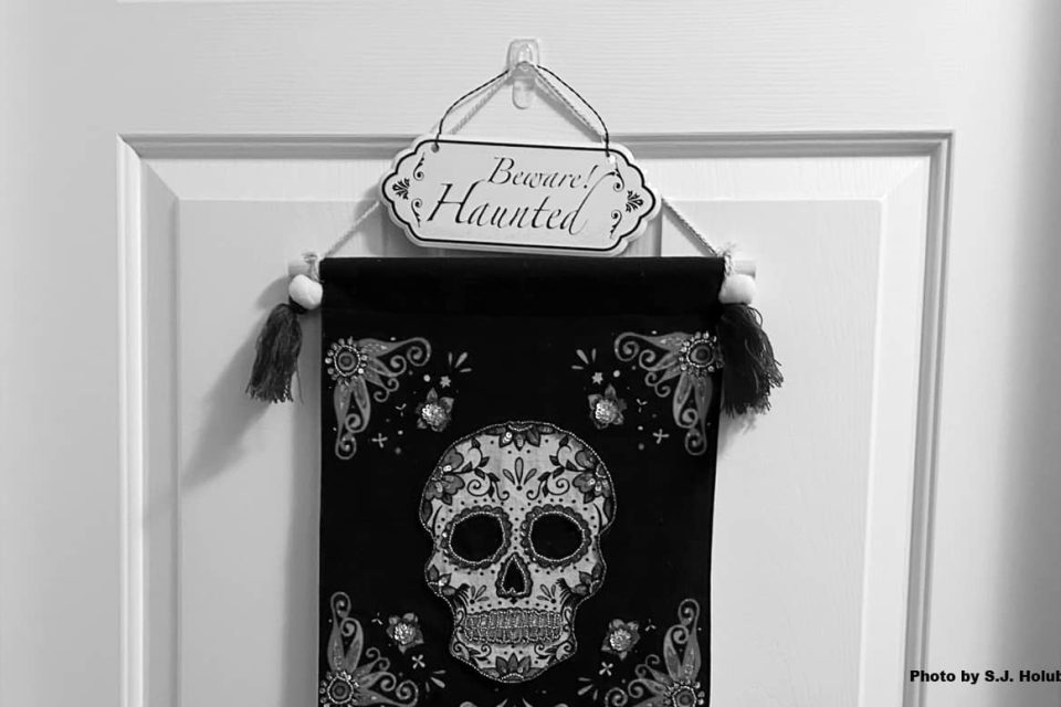 A black and white photo of a white bedroom door with a small metal sign that reads "Beware – Haunted" and a Dia De Los Muertos style fabric wall hanging with a skull