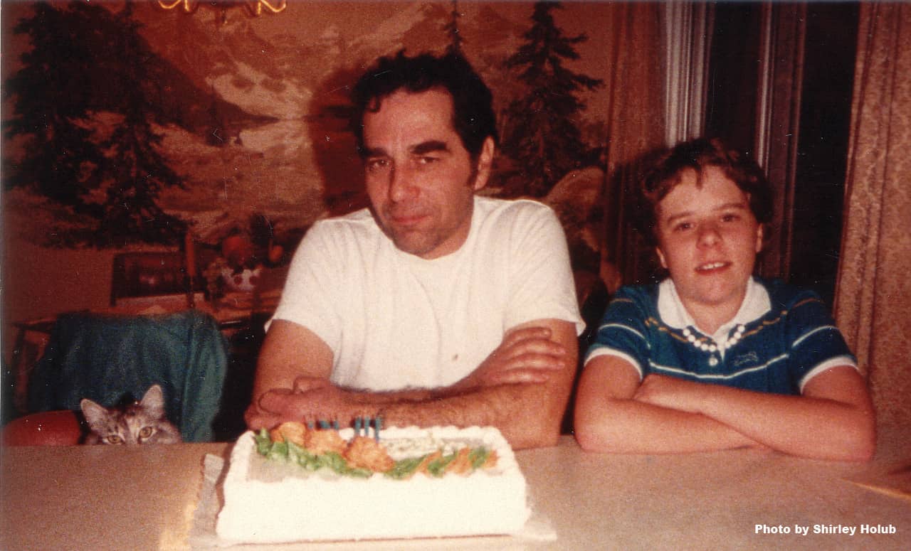 A photo from the 1980s of a dad, a child, and a cat all sitting at a counter with a birthday cake on the counter