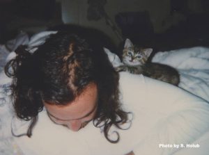 A man with long hair lays on a bed with a tiny tabby kitten on his back