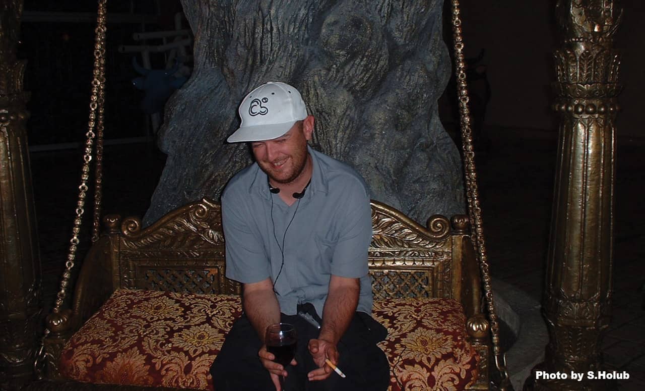 A photo of a man sitting on an ornate swinging bench at a casino in Los Vegas. He is  wearing a white hat and gray shirt and holding a cigarette and a wine glass