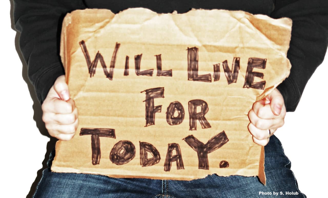A person holds a cardboard sign with the words: will live for today.