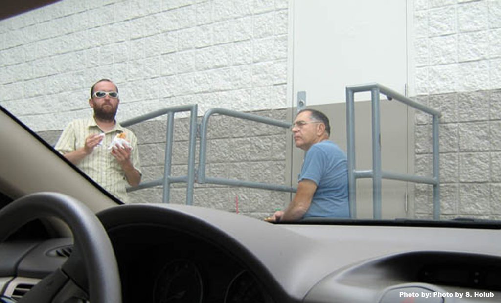 view is from inside a parked car of a man eating a sandwich and another man sitting on some steps