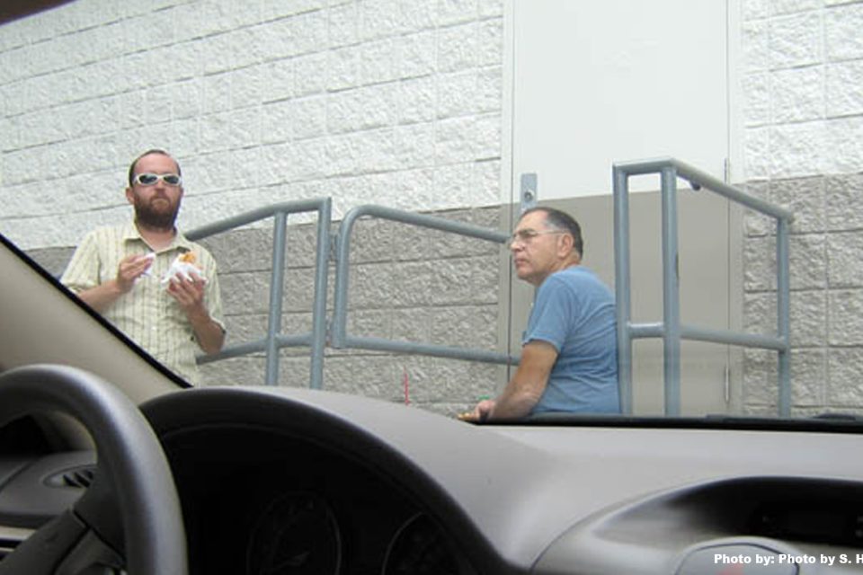 view is from inside a parked car of a man eating a sandwich and another man sitting on some steps